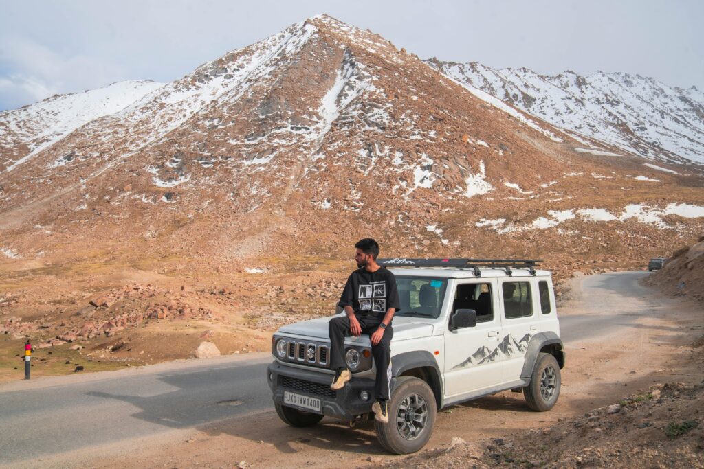 Man sitting on an SUV in the rocky and snowy hills of Ladakh, India, embodies adventure and exploration.